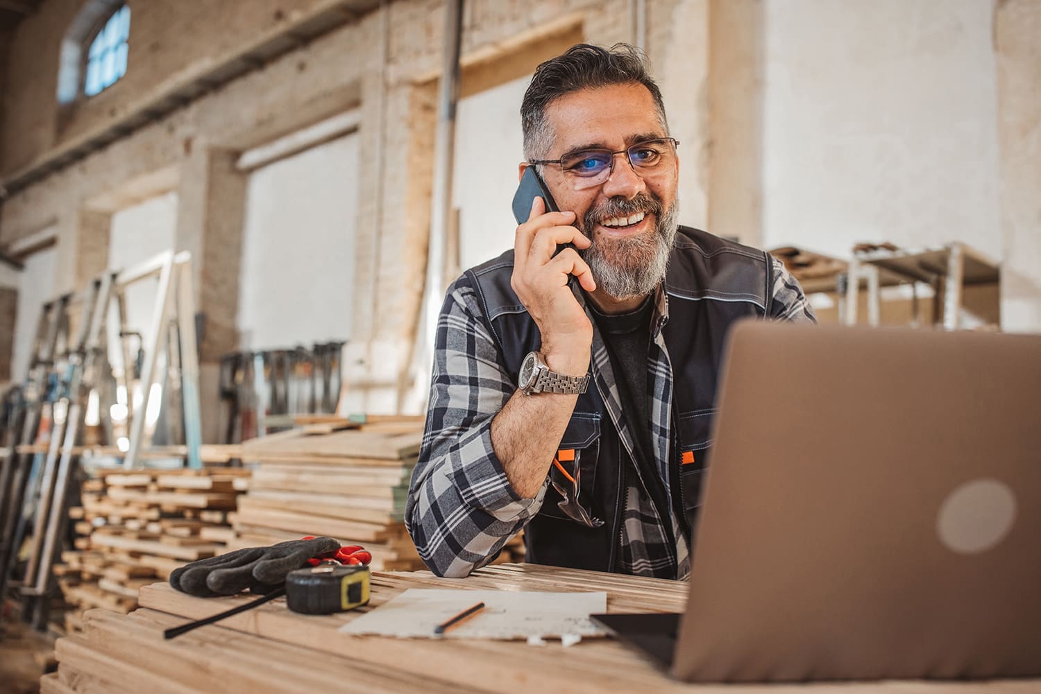 Carpenter in wood workshop Smiling man with glasses and a beard talks on a phone while using a laptop in a workshop filled with wooden planks and tools.