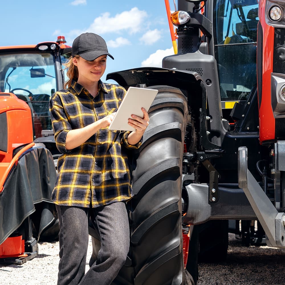 Female sales representative at a dealership for construction and agricultural machinery stands next to a tractor and using digital tablet. A person in a plaid shirt and black cap uses a tablet while leaning against a red tractor on a sunny day. Another tractor is visible in the background.
