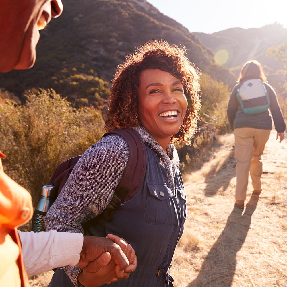Woman Helping Man On Trail As Group Of Senior Friends Go Hiking In Countryside Together Person smiling while holding hands with another person on a hiking trail in a sunny mountainous area. Another person walks ahead.