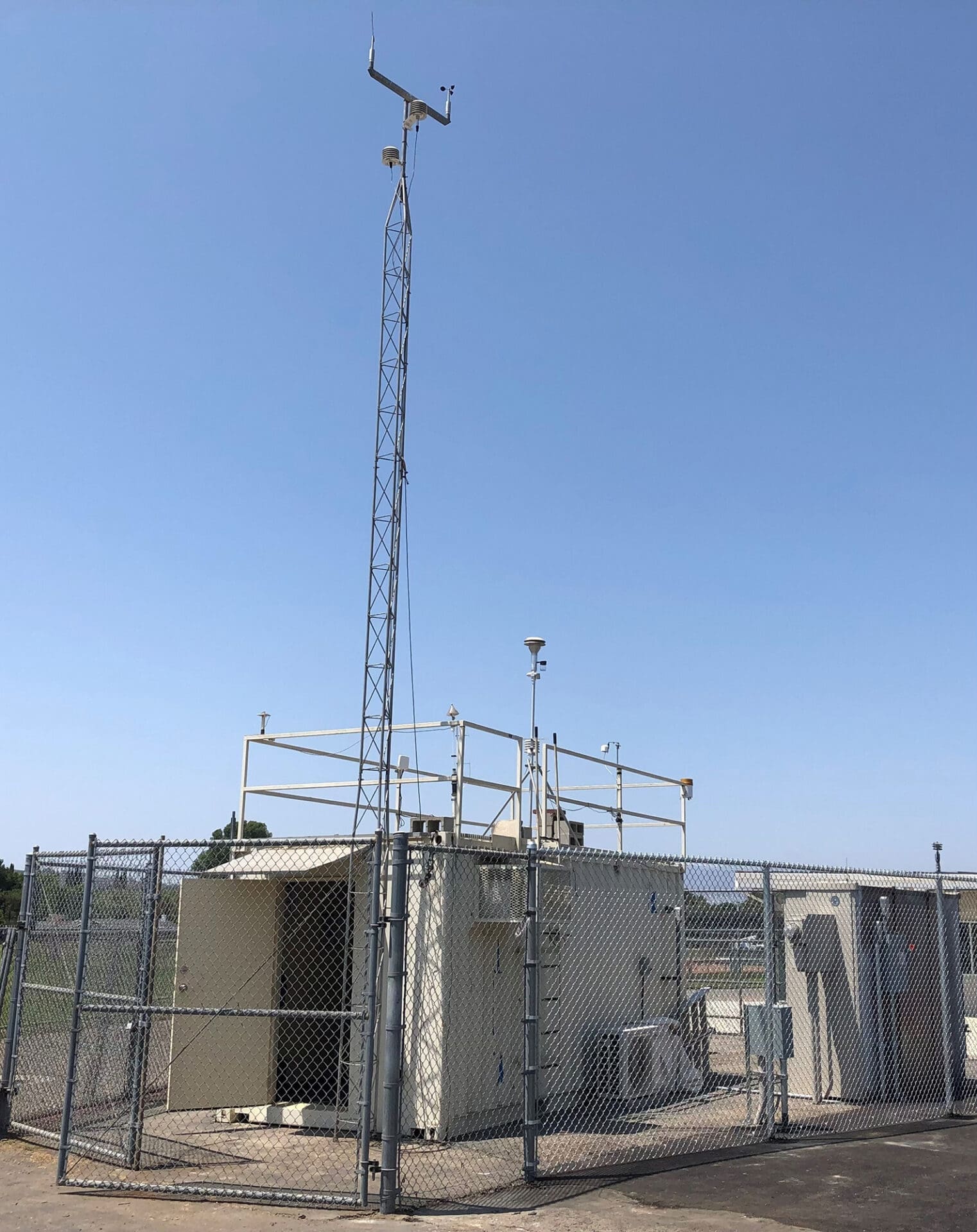Thousand-Oaks-Monitoring-Station-web A fenced weather station with instruments on a rooftop and a tall antenna against a clear blue sky.