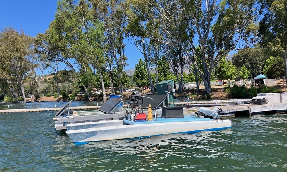Casitas Rowing coaching boat 2-cropped Small pontoon boat docked on a lake with trees and picnic areas in the background under a clear blue sky.