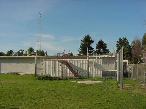 Simi A single-story concrete building with a metal staircase on its roof, surrounded by grass and a chain-link fence. Trees and a clear sky are in the background.