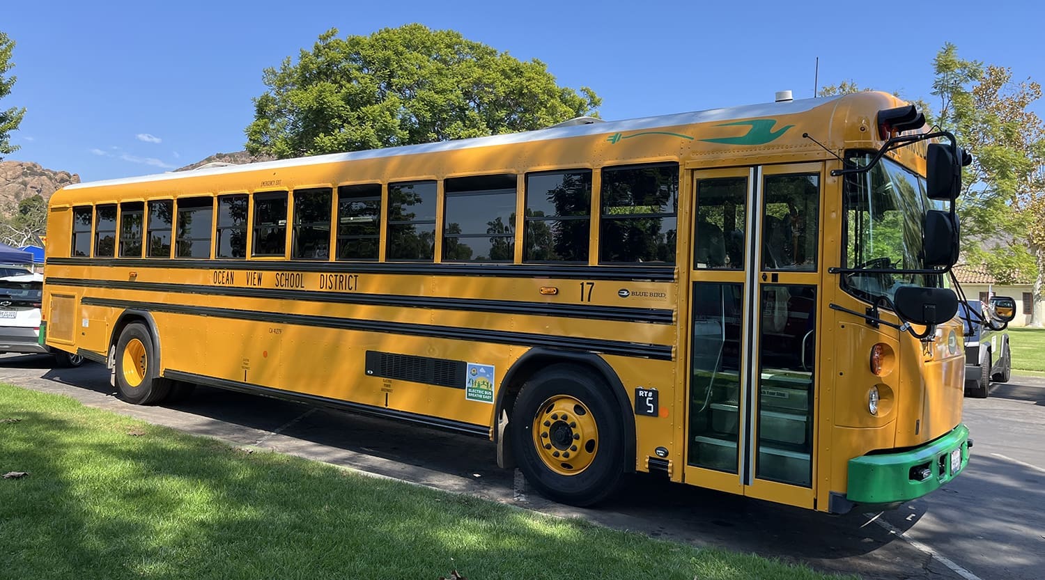 Ocean View School District Electric Bus-web Yellow school bus parked on a driveway, with Ocean View School District written on its side. Trees and a clear blue sky are in the background.