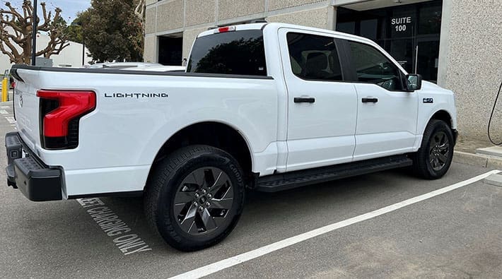 A white electric Ford 150 Lightning truck is parked in a lot outside a building.