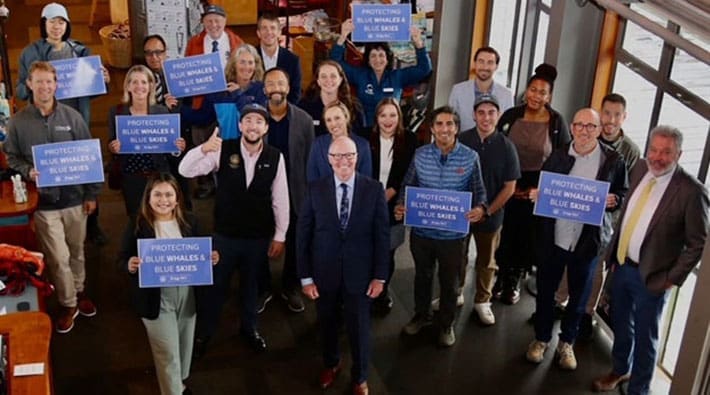 A group of people, some holding signs saying "Protecting Blue Whales and Blue Skies" look up at the camera.