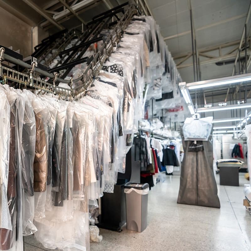 Clothing covered in plastic hangs on a conveyor system in a dry cleaning facility with fluorescent lighting.