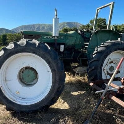 A green tractor with large white wheels is parked on dry grass near a plow attachment. Hills and trees are visible in the background under a clear blue sky.