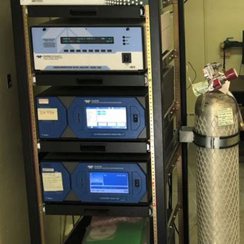 Rack-mounted electronic equipment with a blue interface screen and control panels. A large metal cylinder with a valve is positioned on the right side.