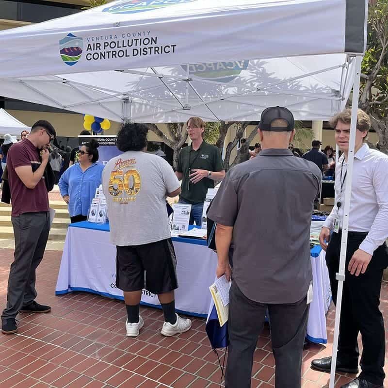 Several people talk at a booth with informational materials under a Ventura County Air Pollution Control District canopy.