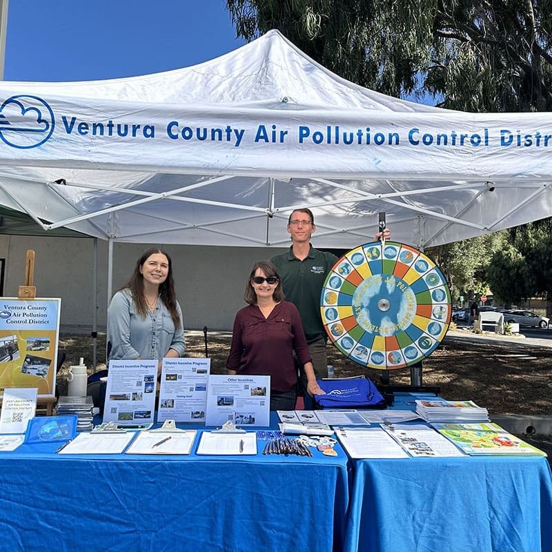 Three people stand at a Ventura County Air Pollution Control District booth with informational materials and a prize wheel under a tent.